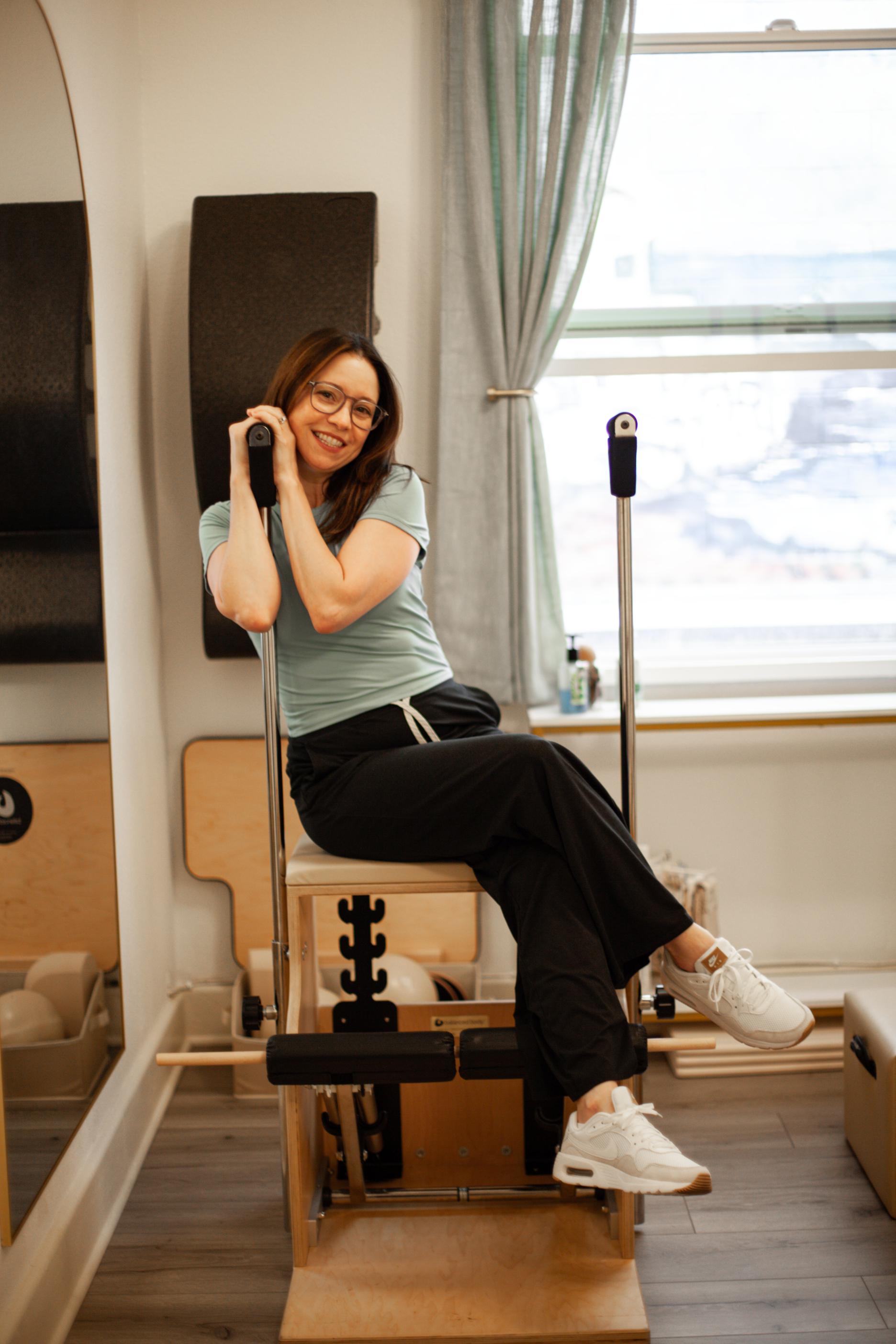Woman sitting on Pilates Combo Chair beside a mirror with a window in the background.
