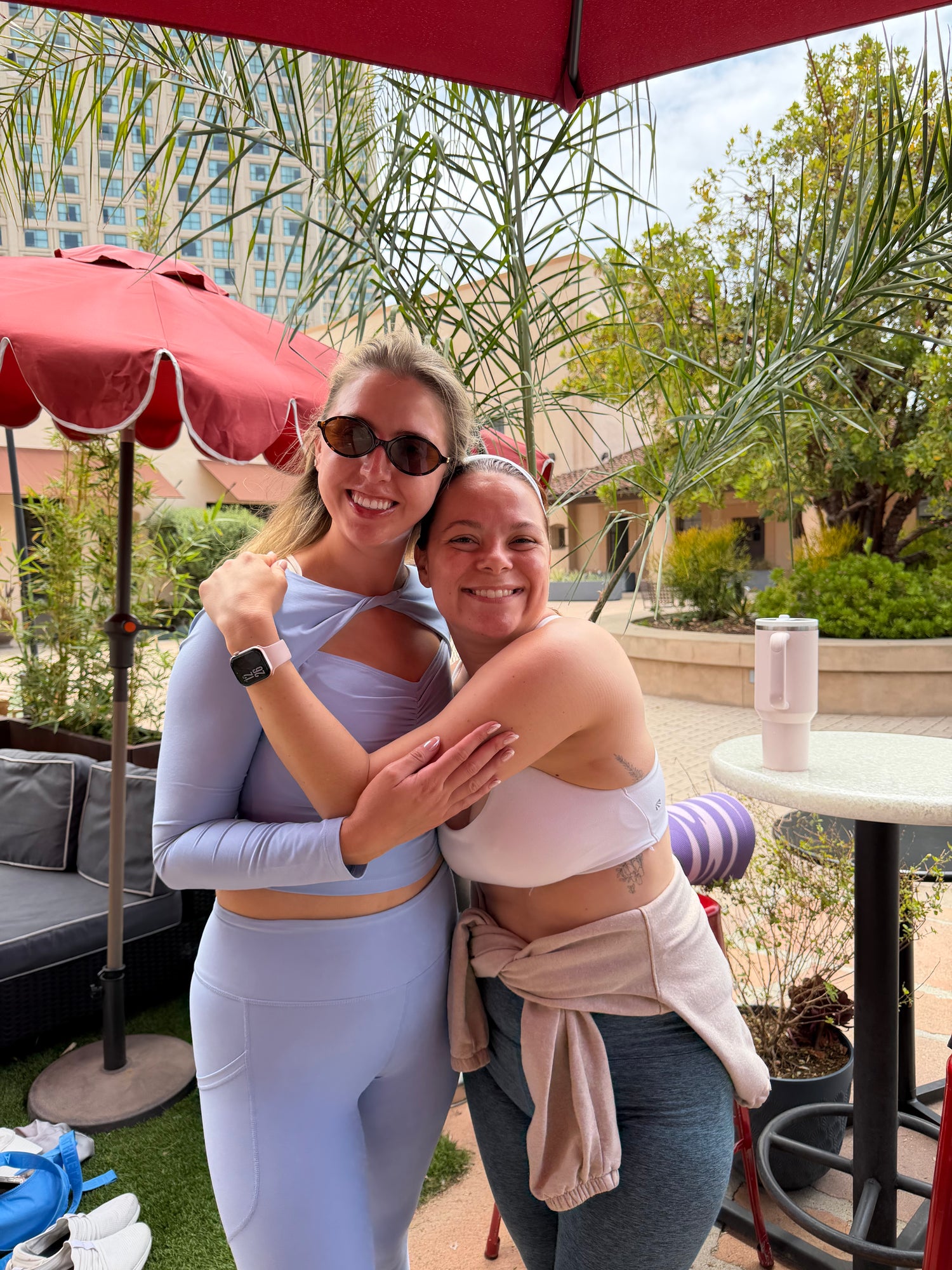 Two women posing together outdoors under a red patio umbrella. The woman on the left wears a light lavender long-sleeve crop top and matching leggings, while the woman on the right wears a pale pink sports bra and a beige shirt tied around her waist.