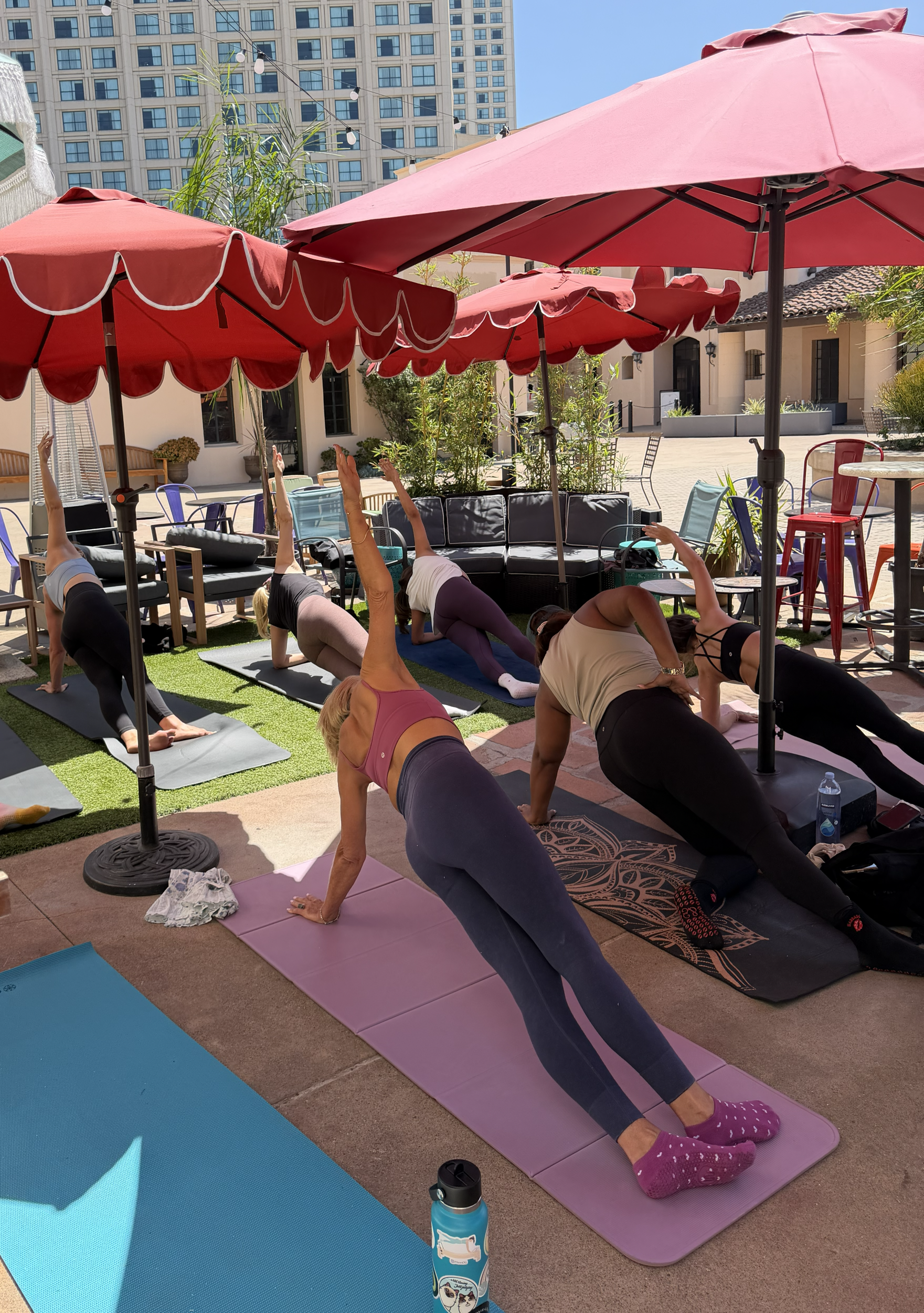 Group of people doing Pilates outdoors under pink umbrellas with a cityscape in the background.