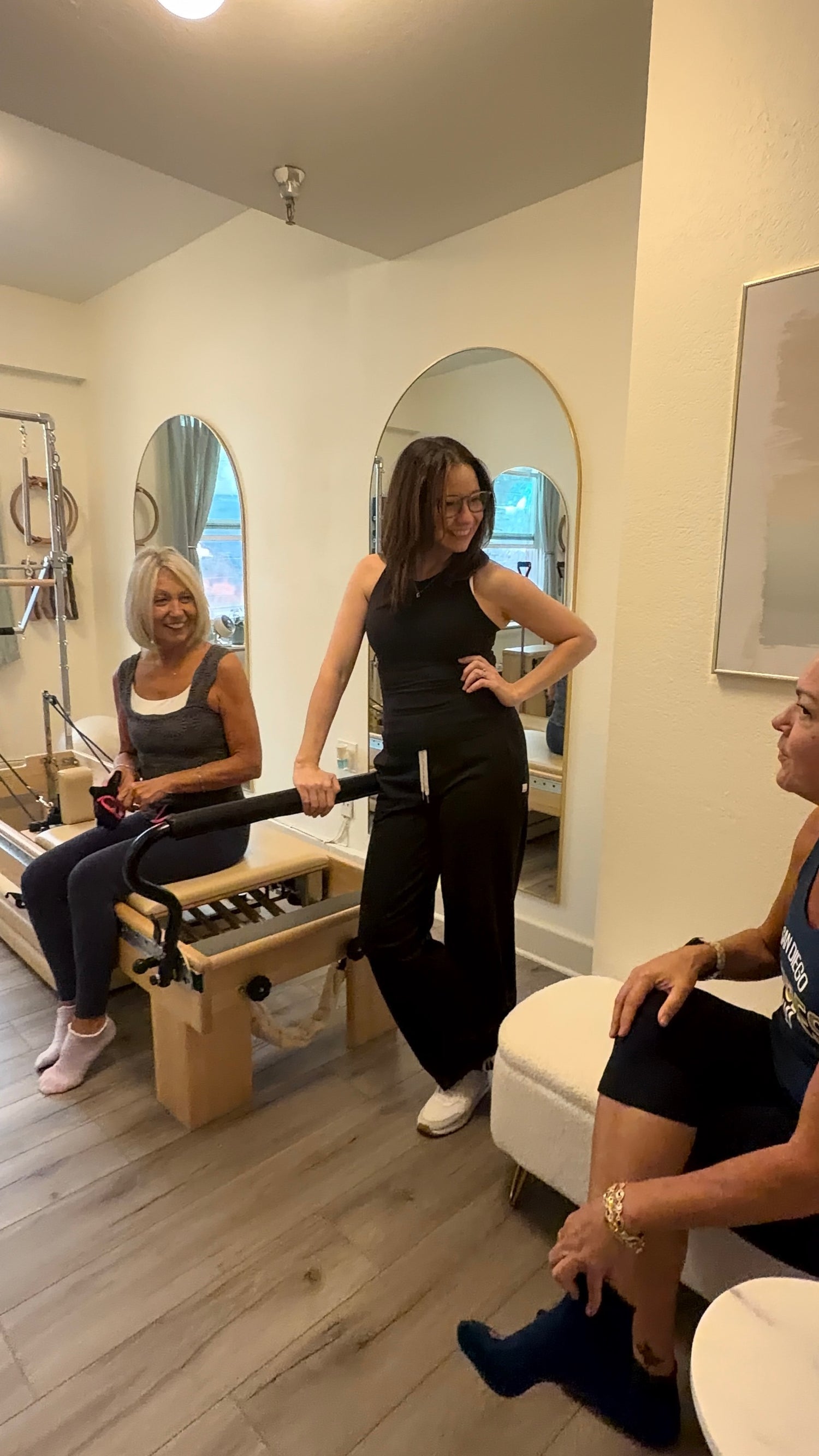 Three women sitting and standing around a Pilates reformer talking.